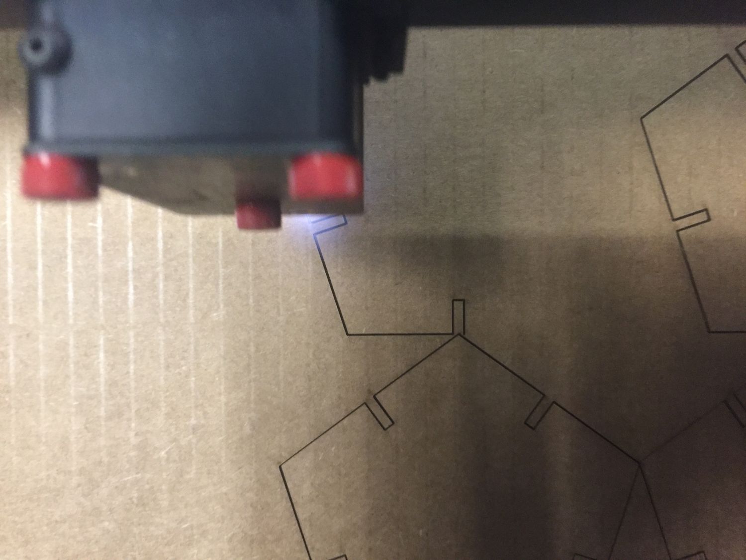 The grey head of laser cutter with red screws is making a line on a piece of cardboard